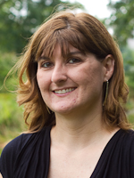 A headshot of a woman looking at the camera. The woman is positioned between a head-on and three-quarters view. Her face and head are clearly visible without sunglasses or a hat. She appears to be outdoors and trees and foliage are visible in the background.